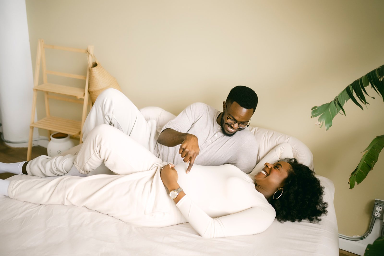 A man and woman are smiling and laughing on a bed in a cozy room, with the woman lying on her side and the man sitting beside her, playfully touching her stomach.