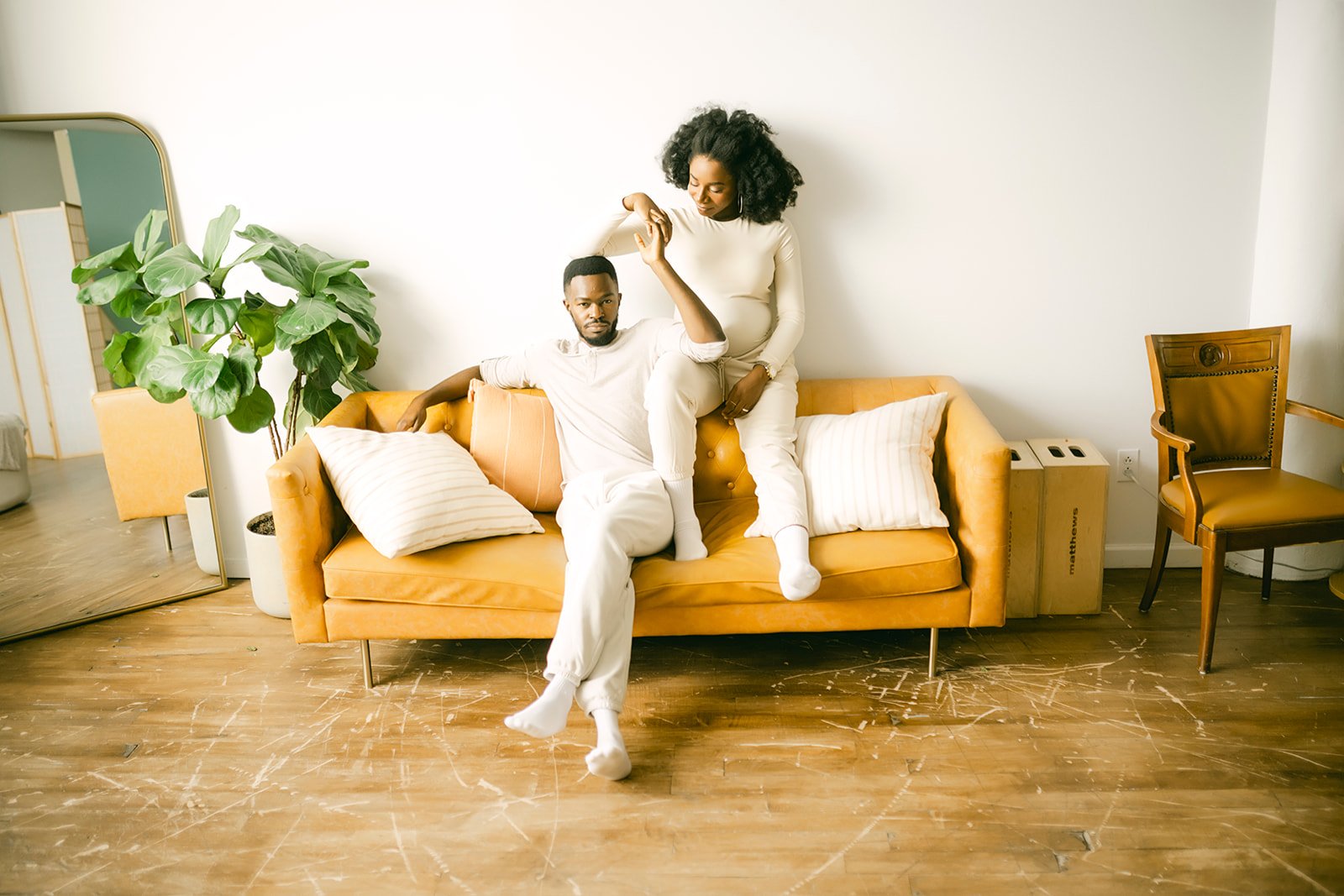 A man and woman with dark curly hair sitting on a mustard-colored sofa in a minimalistic room. The man is sitting with his legs crossed, wearing a light-colored outfit, and the woman is sitting on his lap, looking at him, wearing a matching light-colored outfit.