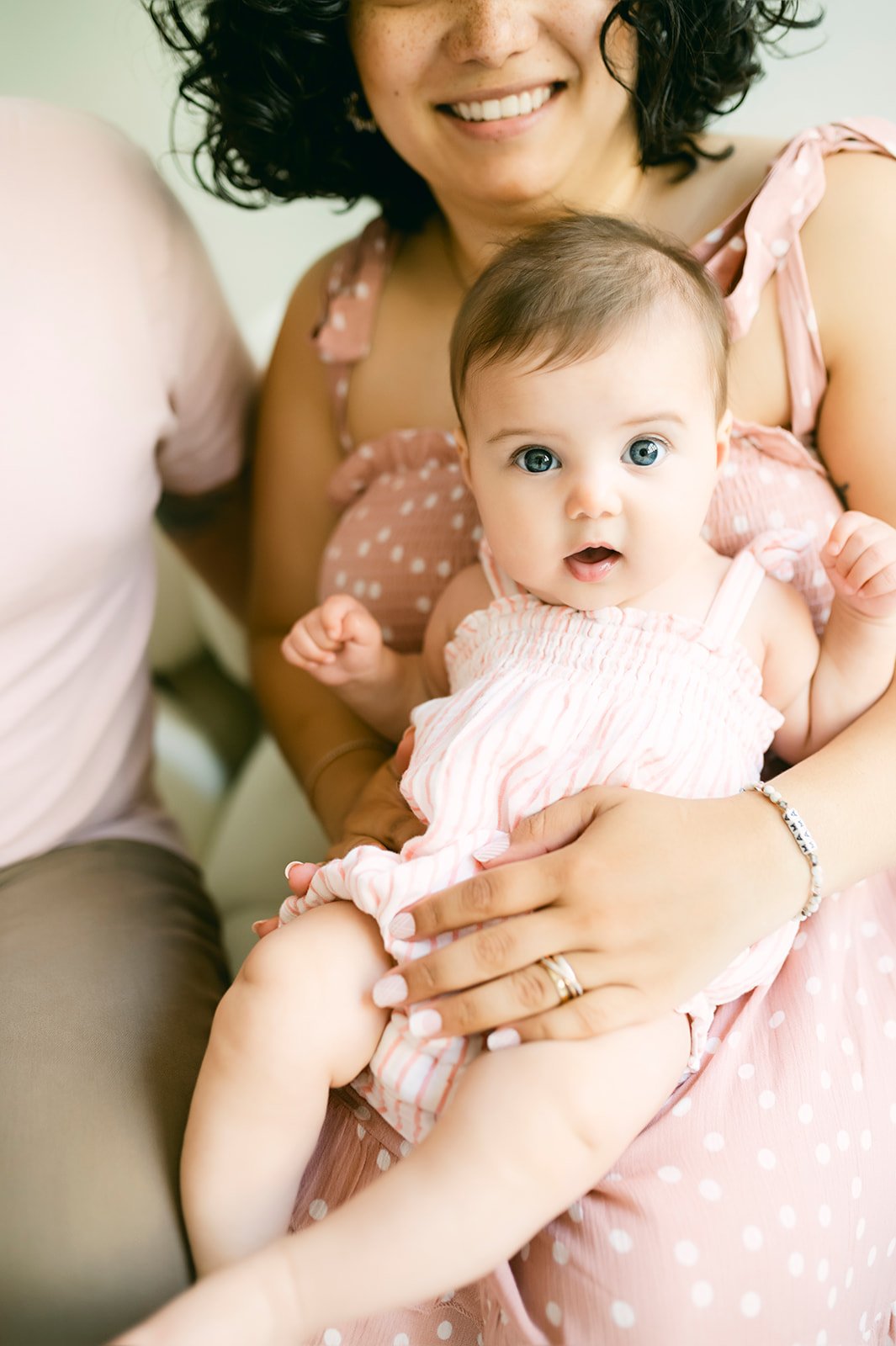 A woman holding a surprised baby girl with big blue eyes, wearing a pink striped dress. The woman has curly dark hair and is smiling.