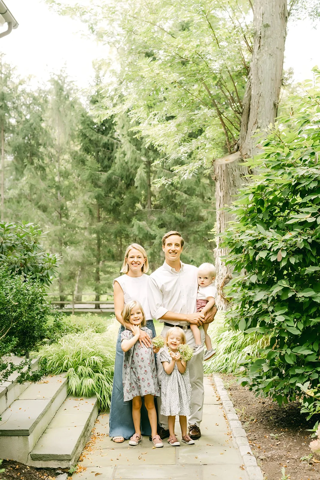 A family of five, including two adults and three children, standing on a garden path surrounded by lush green trees and shrubs.