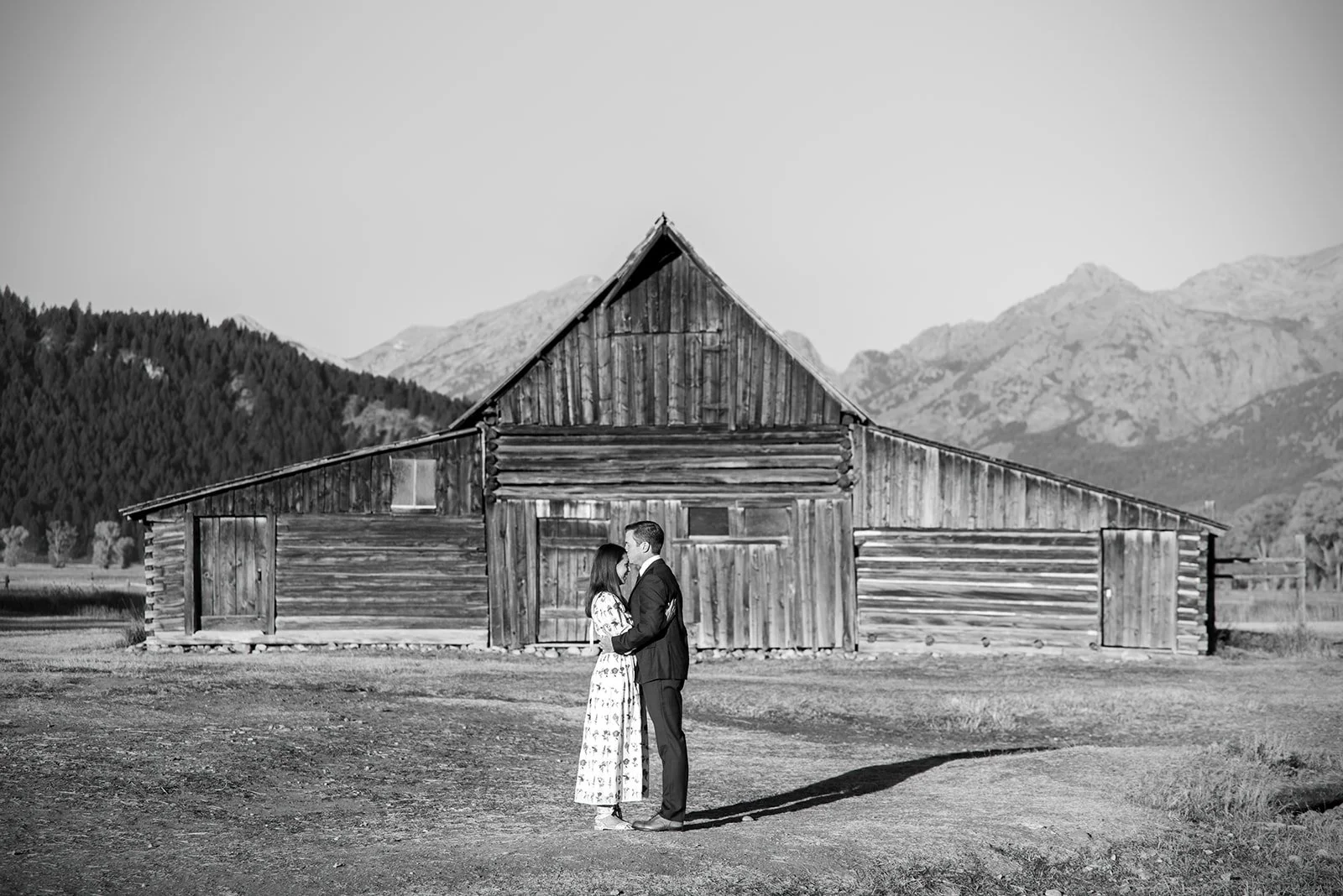 A black and white photo of a couple standing close together, facing each other, holding hands, in front of a rustic wooden barn with mountain scenery in the background.