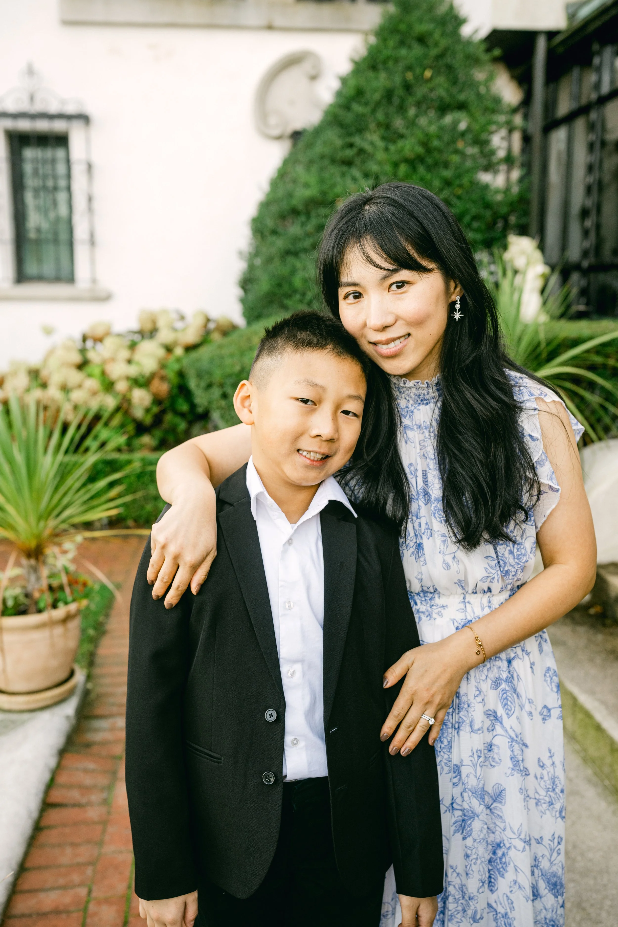 A woman and a boy posing outdoors in front of a house with greenery, smiling, with the woman’s arm around the boy.