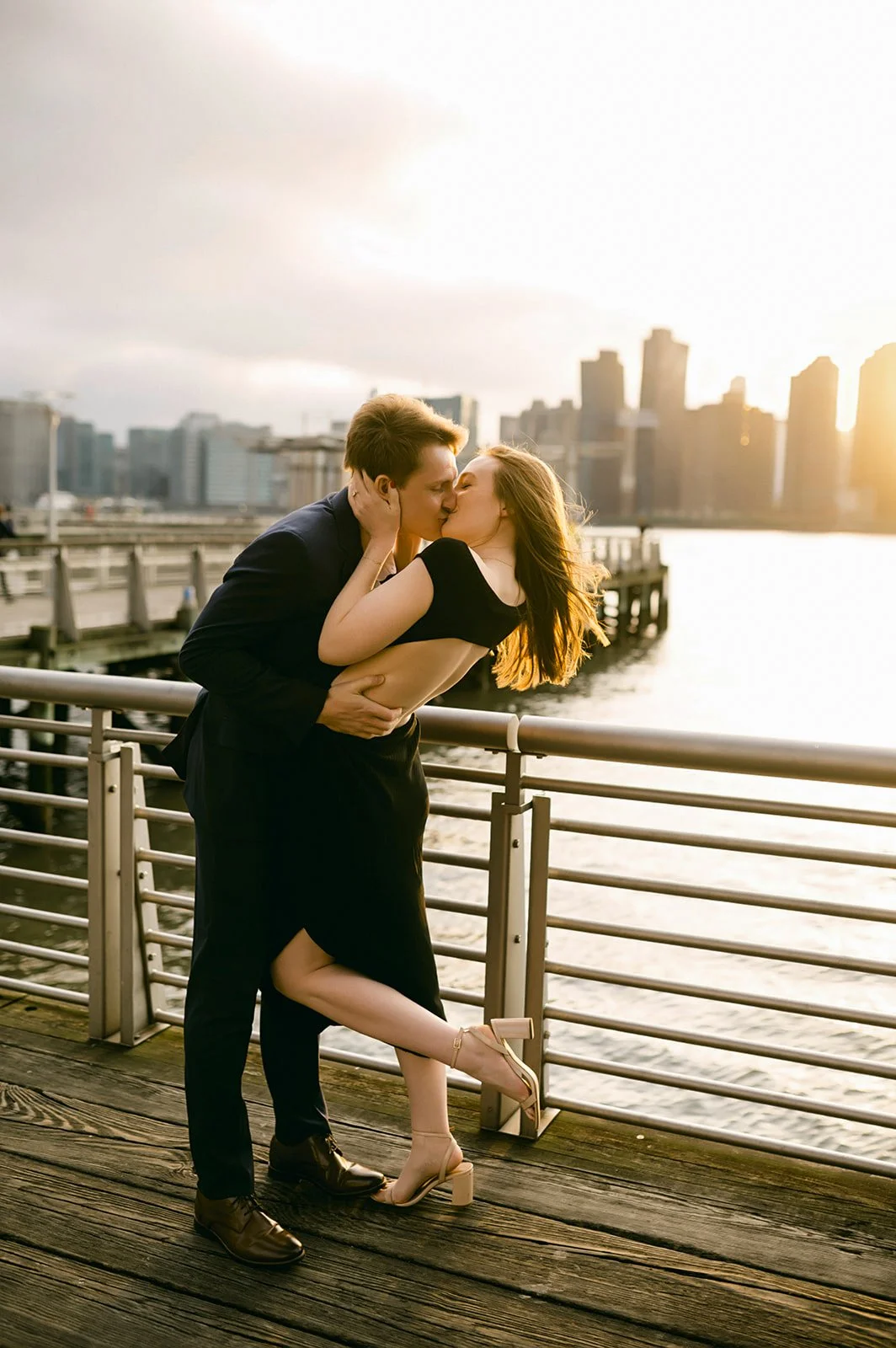 A couple sharing a kiss on a waterfront pier during sunset, with a city skyline in the background.