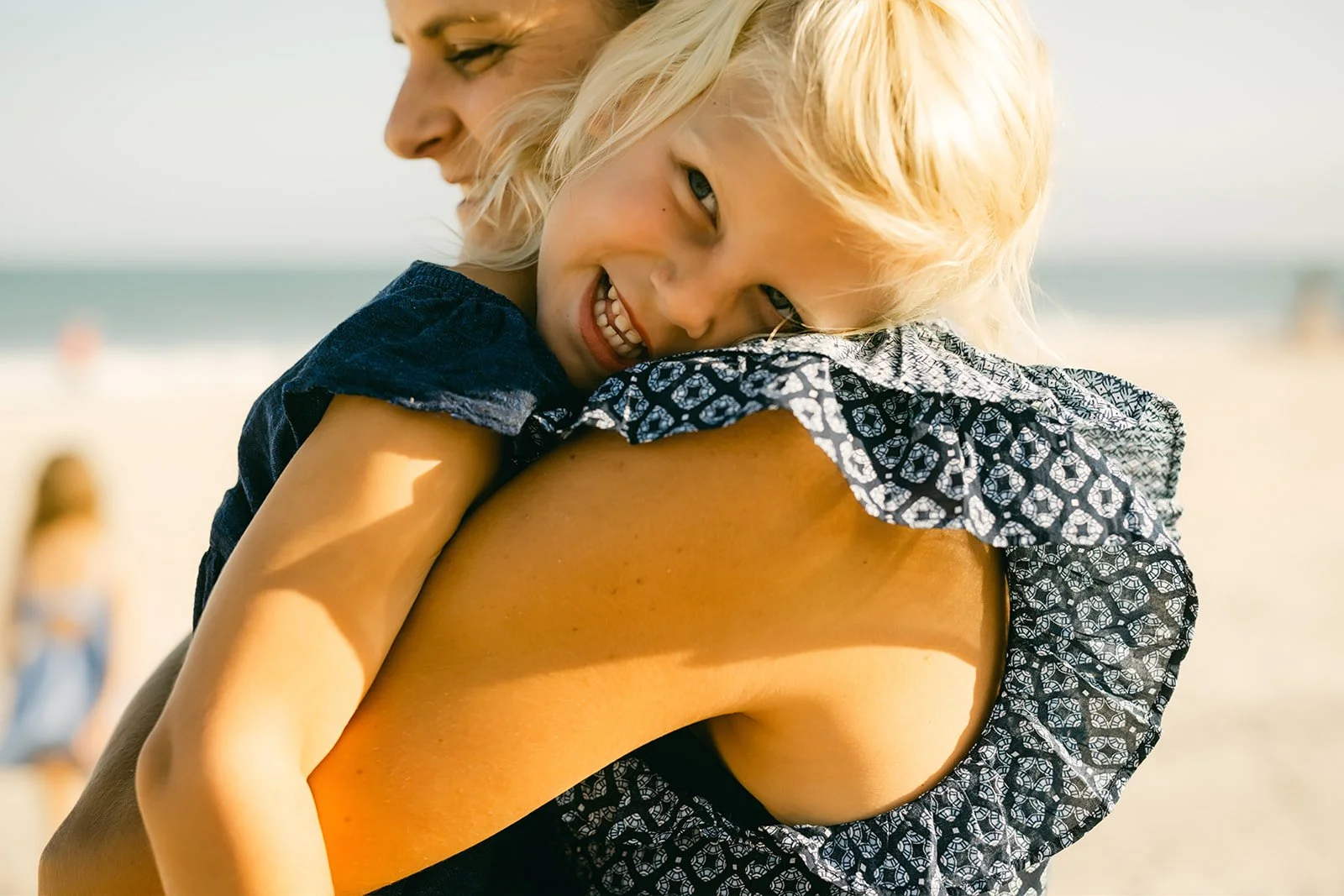 A woman holding a young girl at the beach, smiling, with sand and ocean in the background.