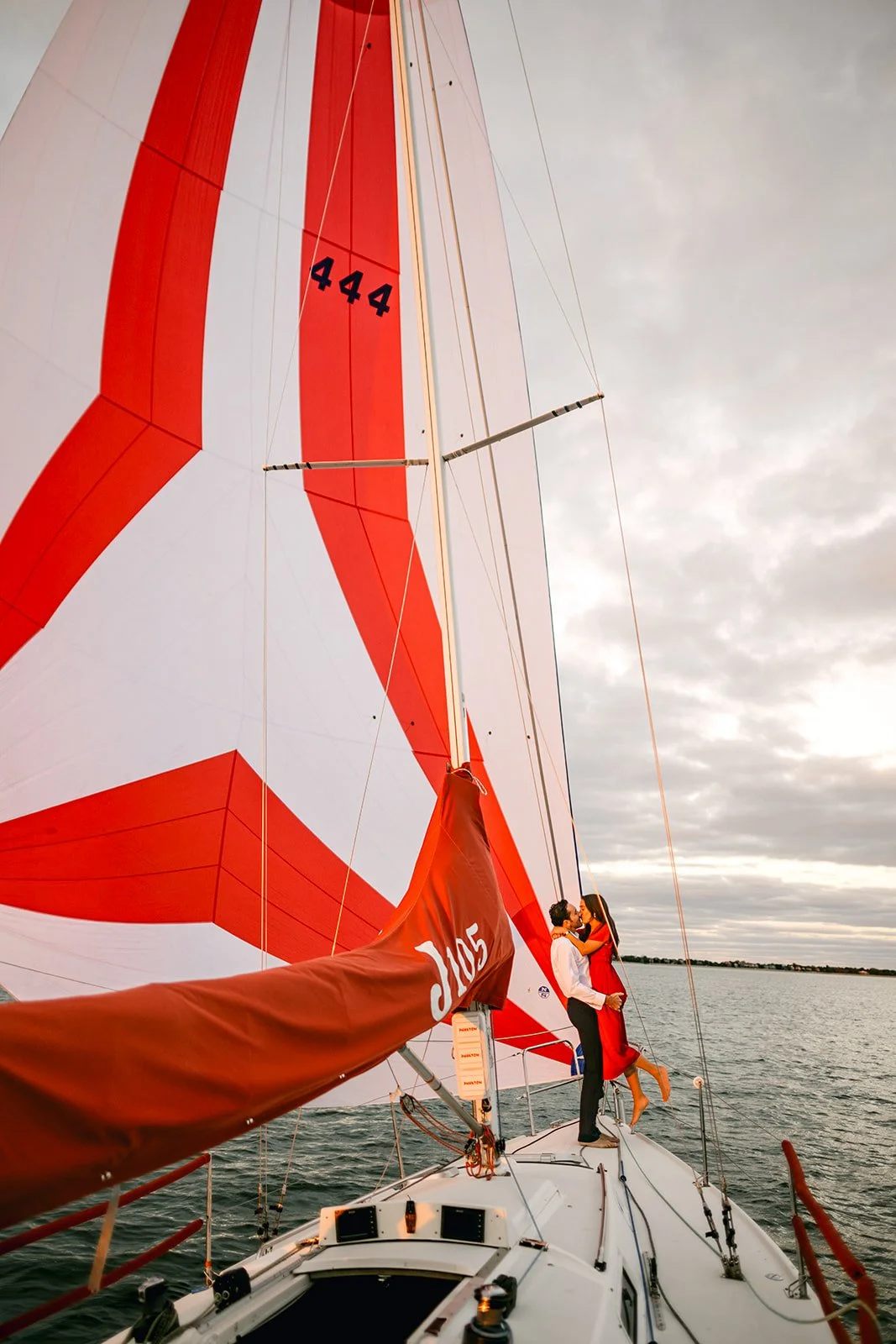 A couple on a sailboat, standing close and embracing, with the man in white shirt and dark pants, the woman in a red dress, on a body of water under a cloudy sky.