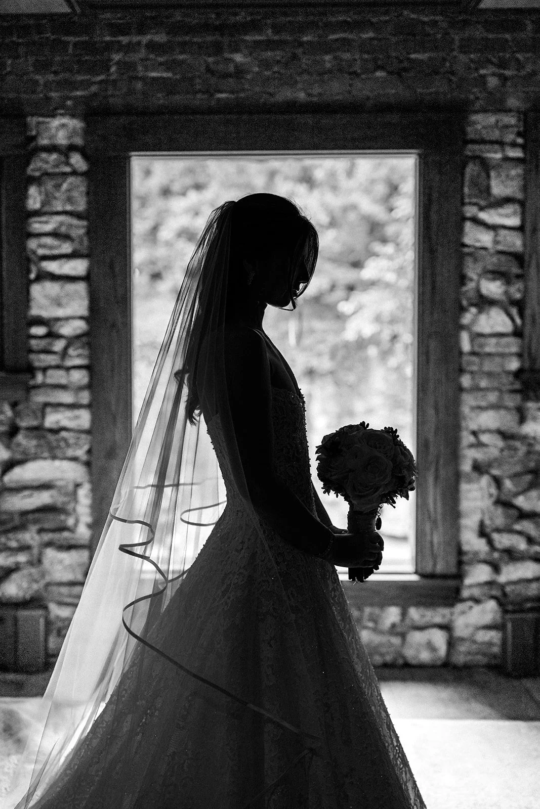 Silhouette of a bride holding a bouquet, standing in front of a window with stone framing.