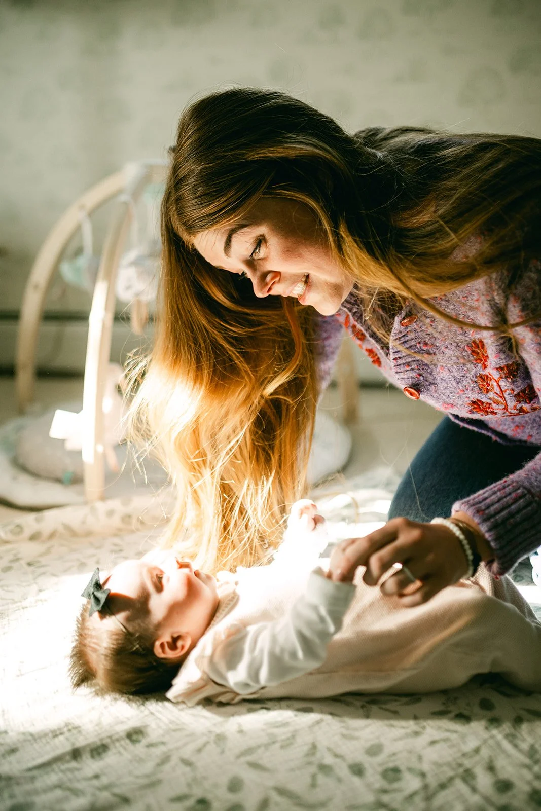 A woman and a baby girl playing together on a bed, with natural light shining on them.