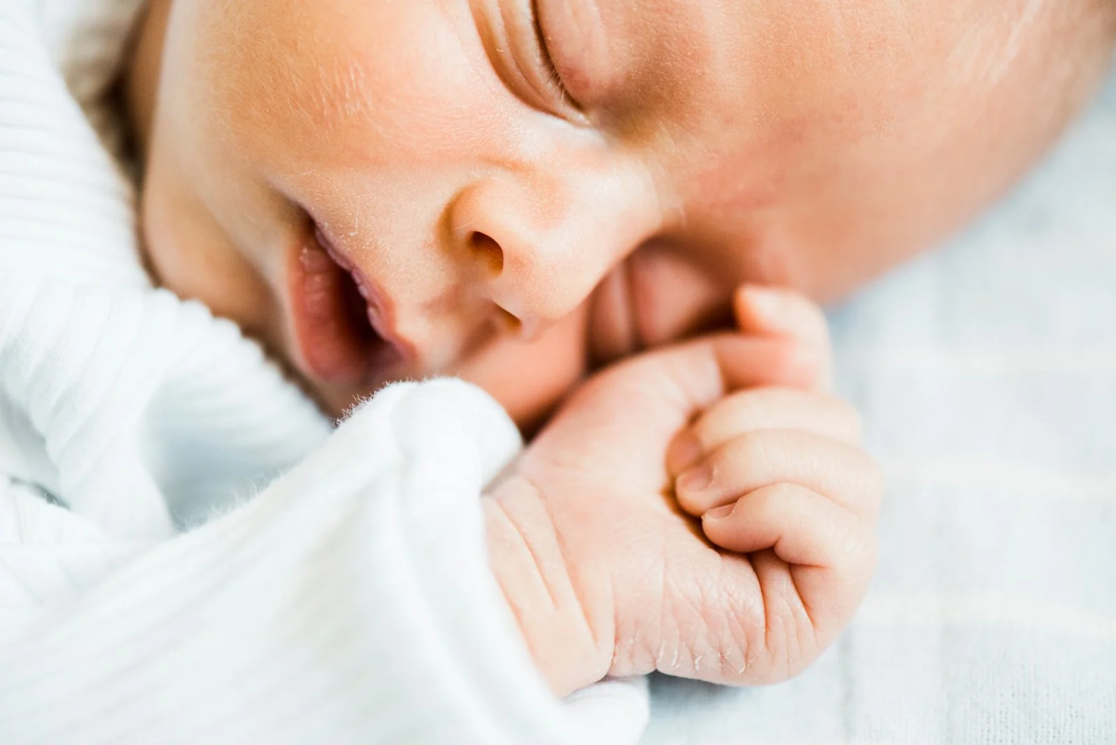 Close-up of a sleeping newborn baby with hand near face, wearing white clothing.