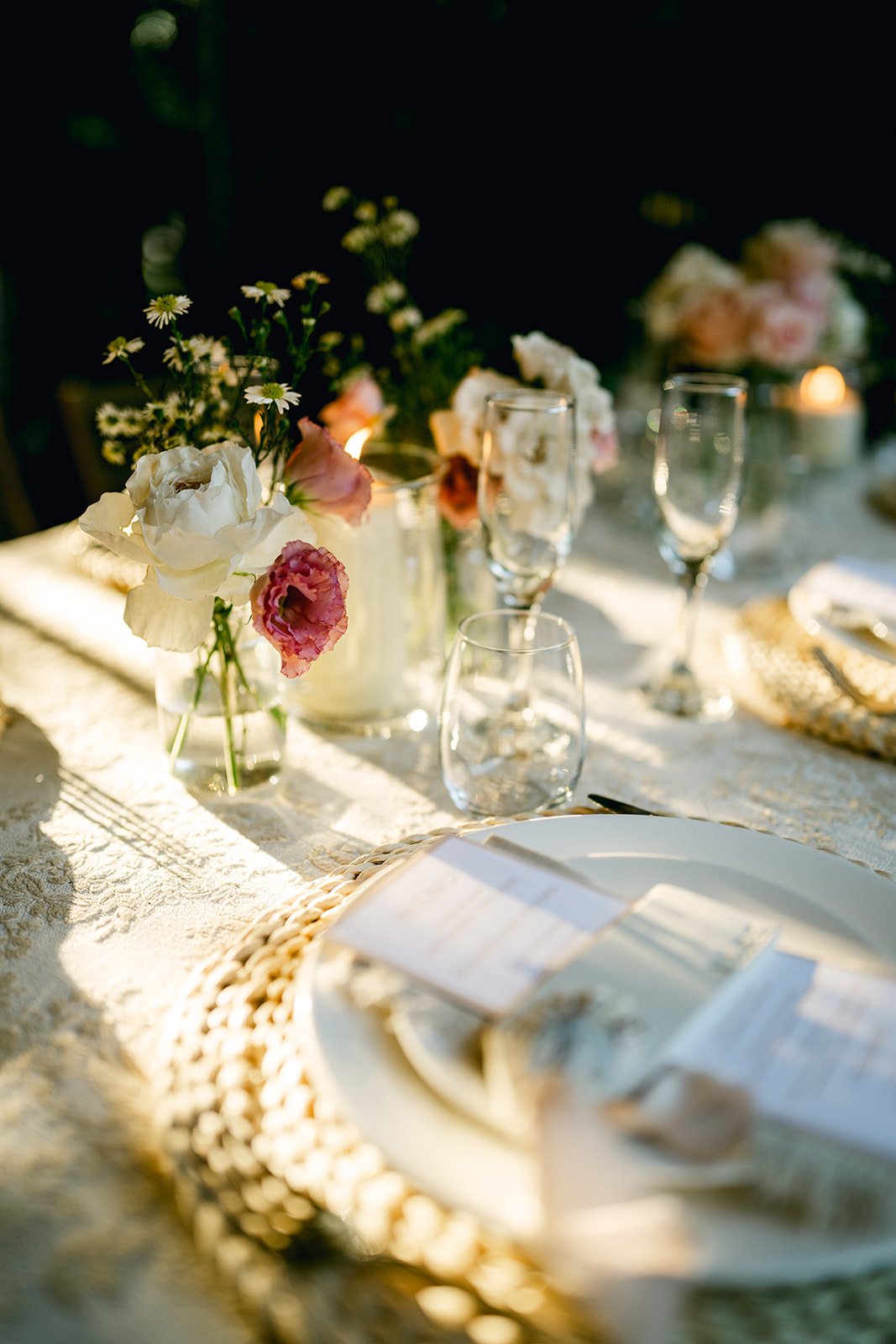 Elegant table setup with floral centerpieces, glassware, and candles, illuminated by warm sunlight.