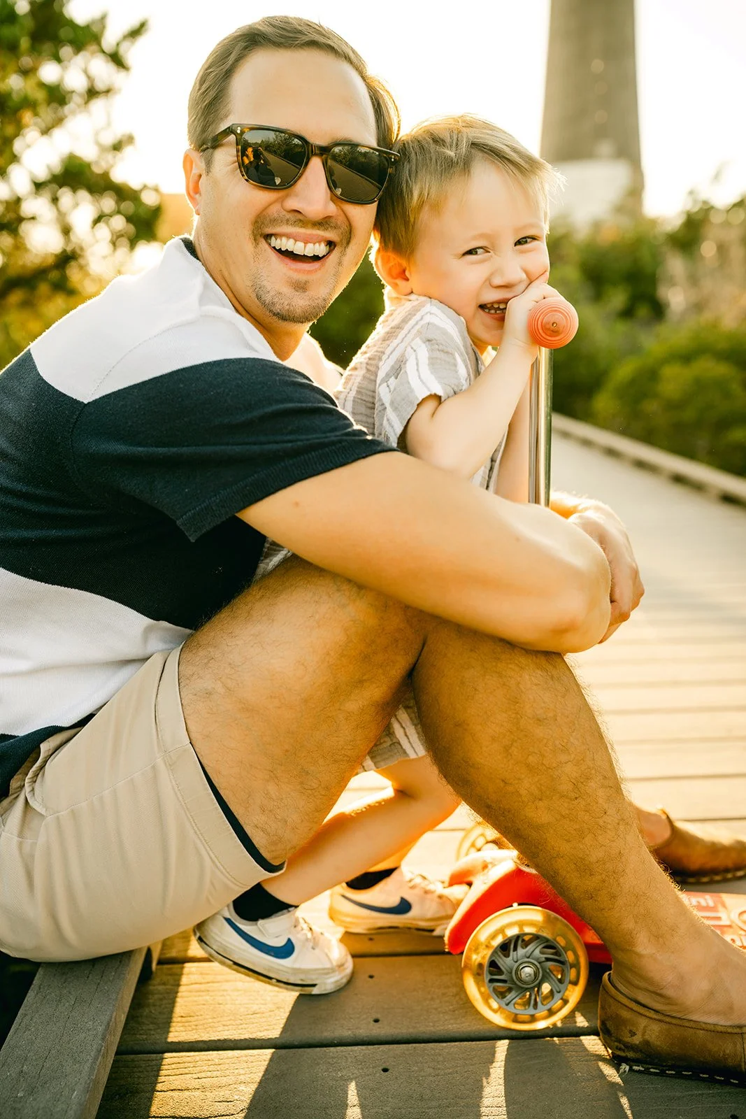 A smiling man wearing sunglasses and a black and white shirt is sitting on a wooden deck, holding a young boy with light brown hair who is laughing and gripping a stick with an orange handle. The boy is seated on the man's knee and has a red skateboard with yellow wheels beside him. The scene is outdoors during sunset with greenery and a tower in the background.