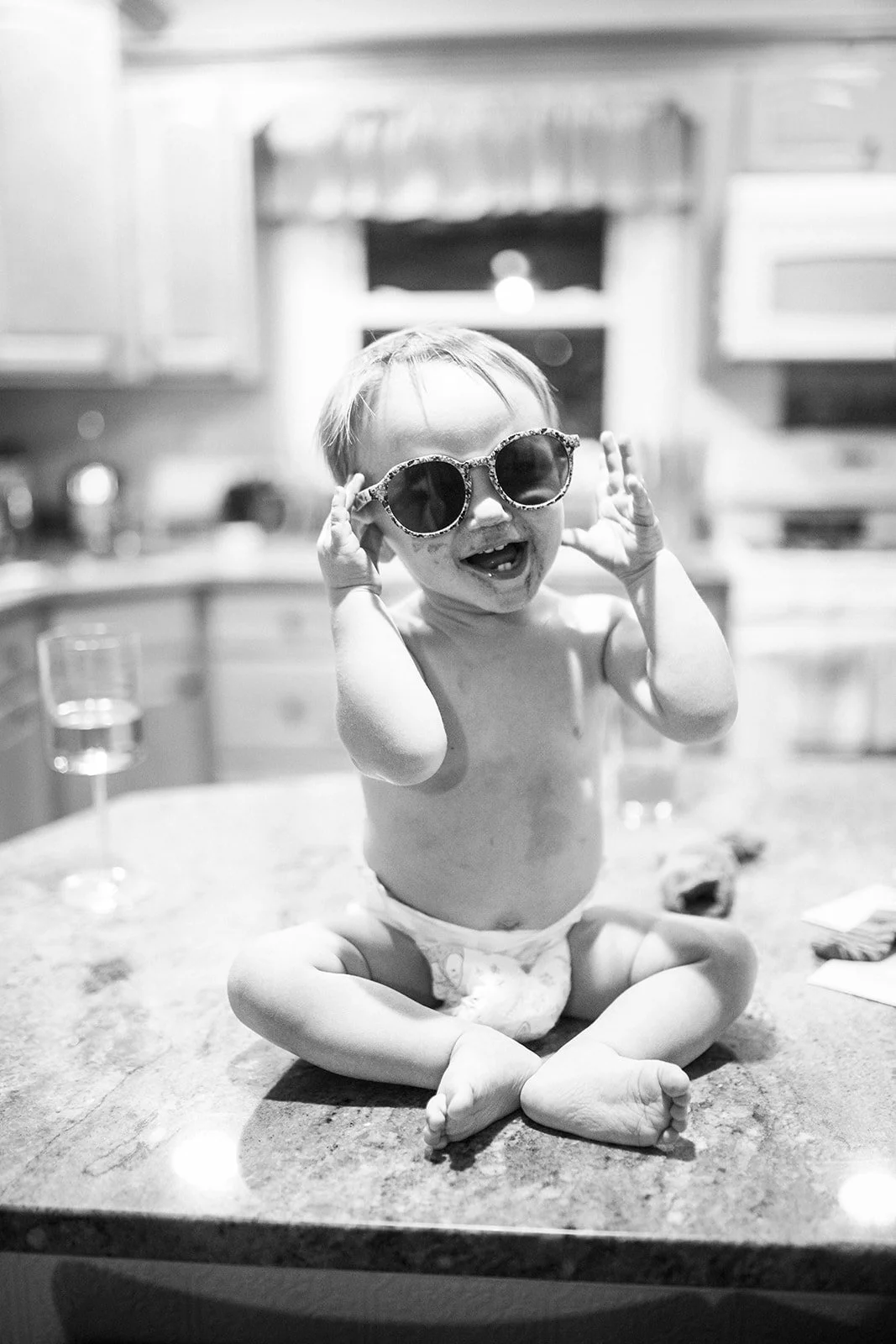 A young child sitting on a kitchen countertop, wearing sunglasses, smiling, and holding the sunglasses with both hands.