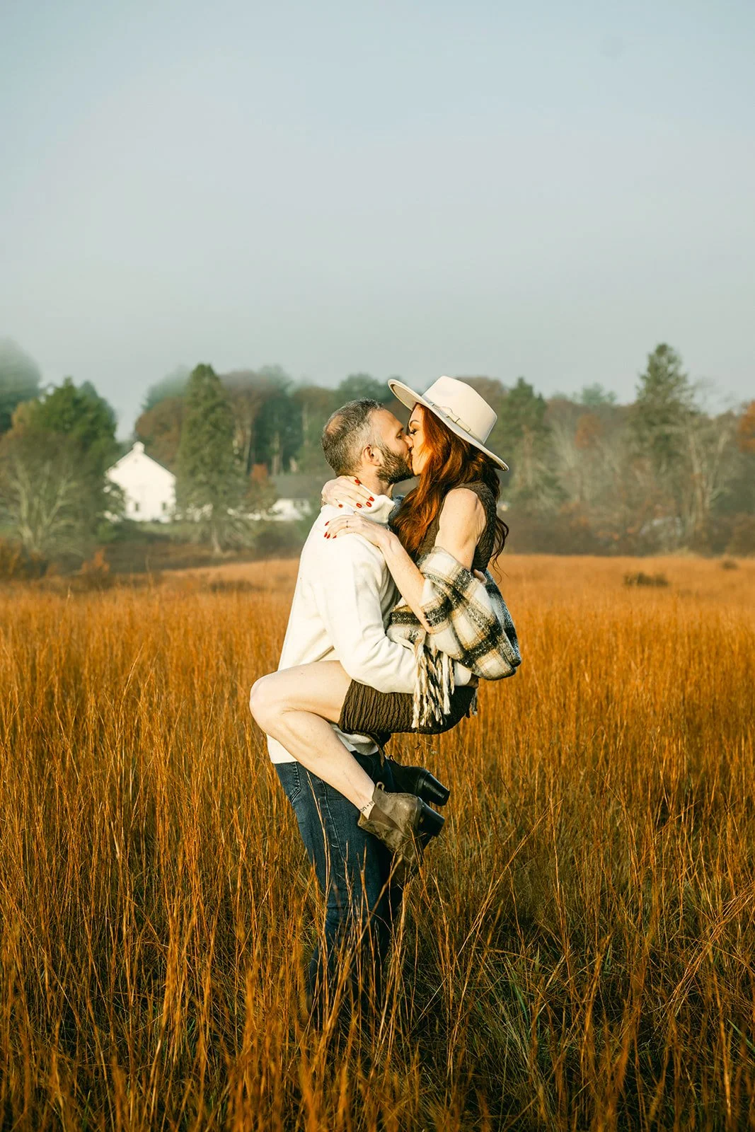 A man lifting a woman in a field of tall, reddish-brown grass, as they share a kiss. The woman wears a wide-brimmed white hat, a plaid shawl, and a dark skirt. The man is dressed in a white jacket and jeans. Trees and houses are visible in the background under a clear sky.