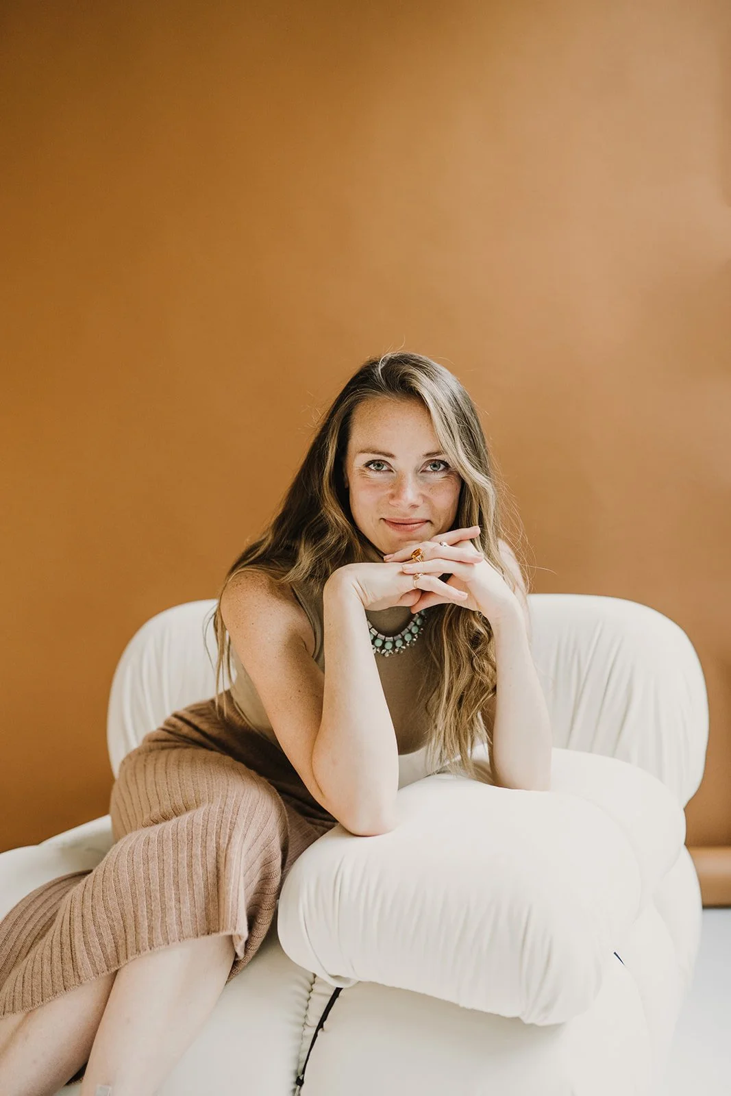 A woman with long wavy hair sitting on a white sofa, leaning forward with her hands clasped under her chin, looking at the camera.