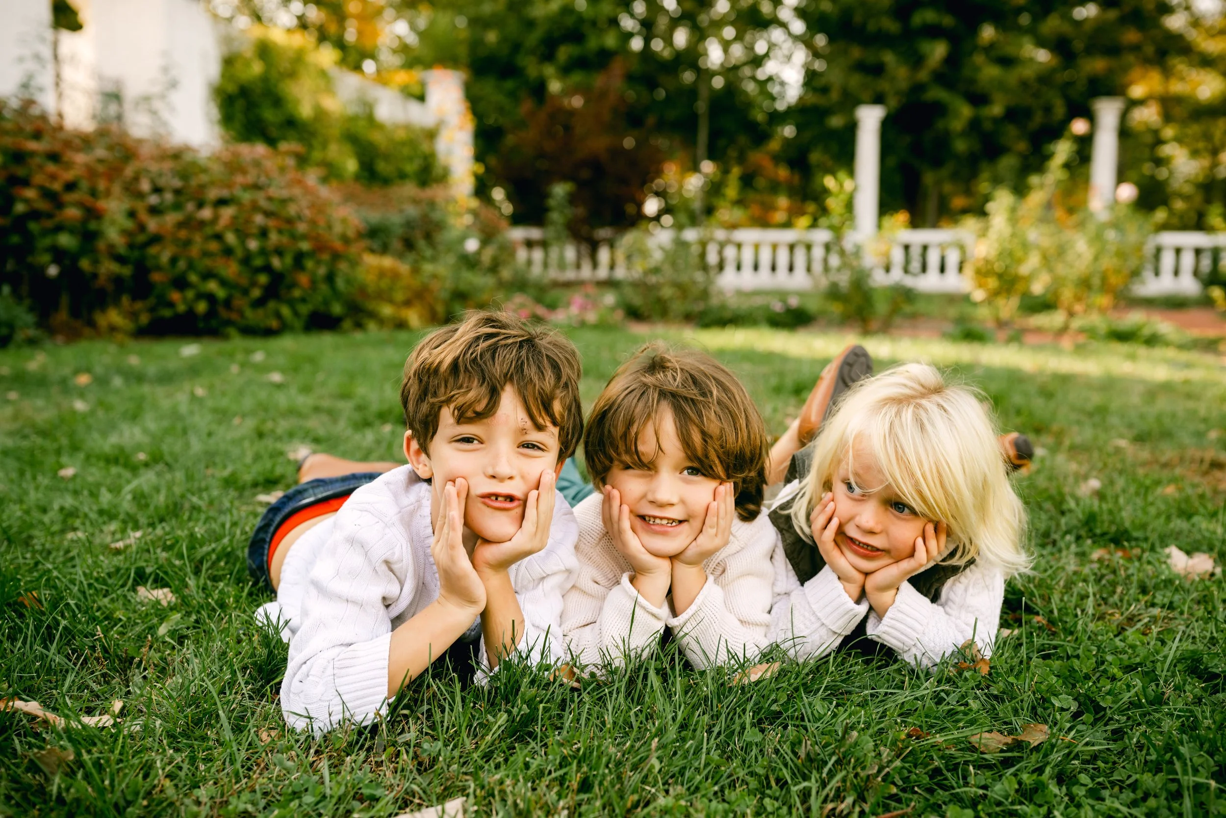 Three children lying on grass in a park, propping up their heads with hands, smiling, with trees and a white fence in the background.