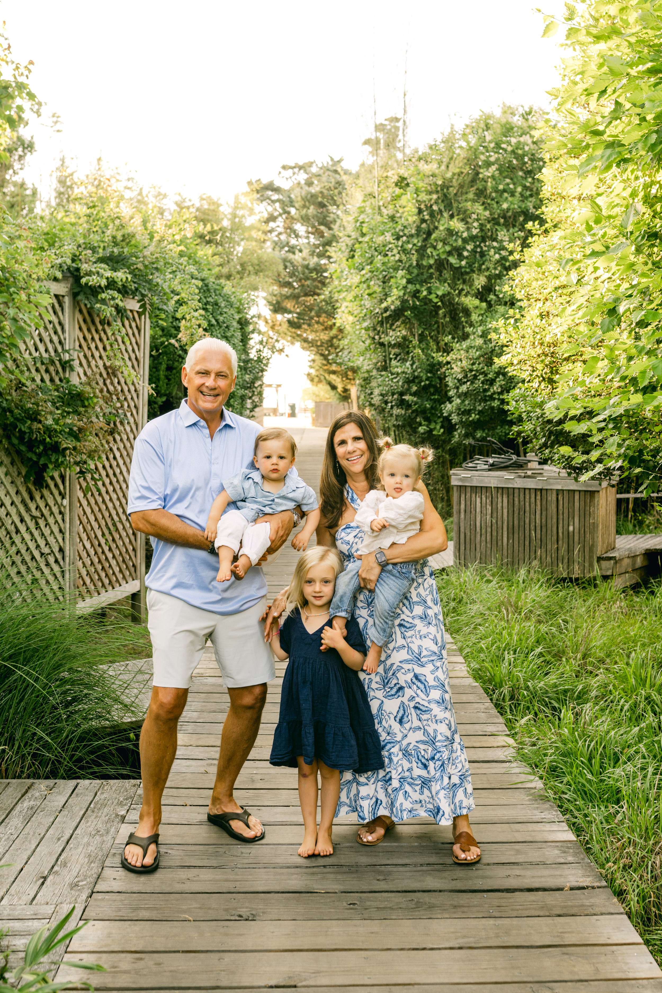 Family of five, including two elderly adults, two young girls, and a young boy, standing on a wooden pathway in a yard with dense green bushes and trees, smiling at the camera during daytime.