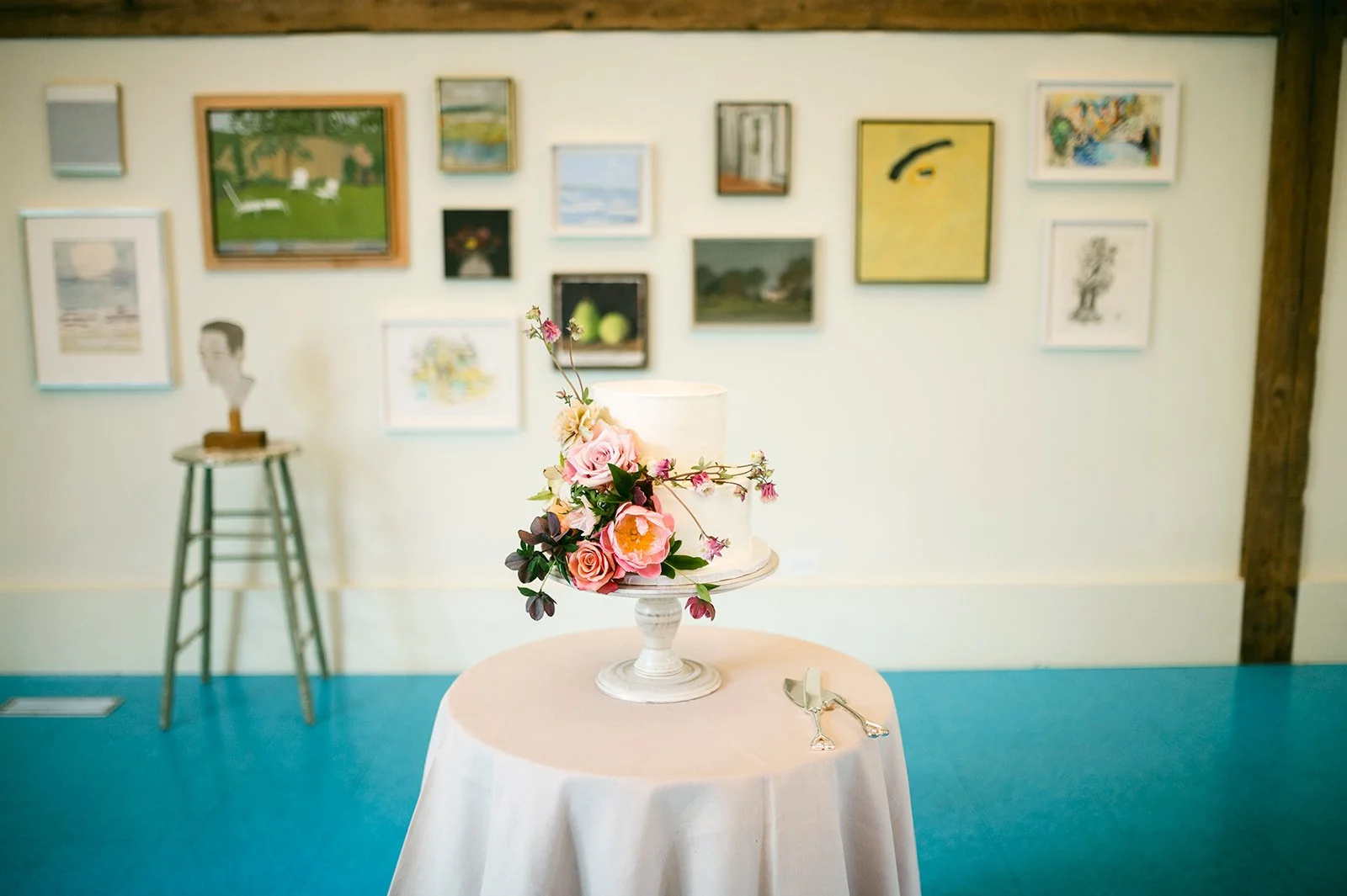 A white wedding cake decorated with pink, cream, and dark purple flowers on a white cake stand, set on a round table with a white tablecloth, located in an art gallery with green and yellow walls and framed paintings.
