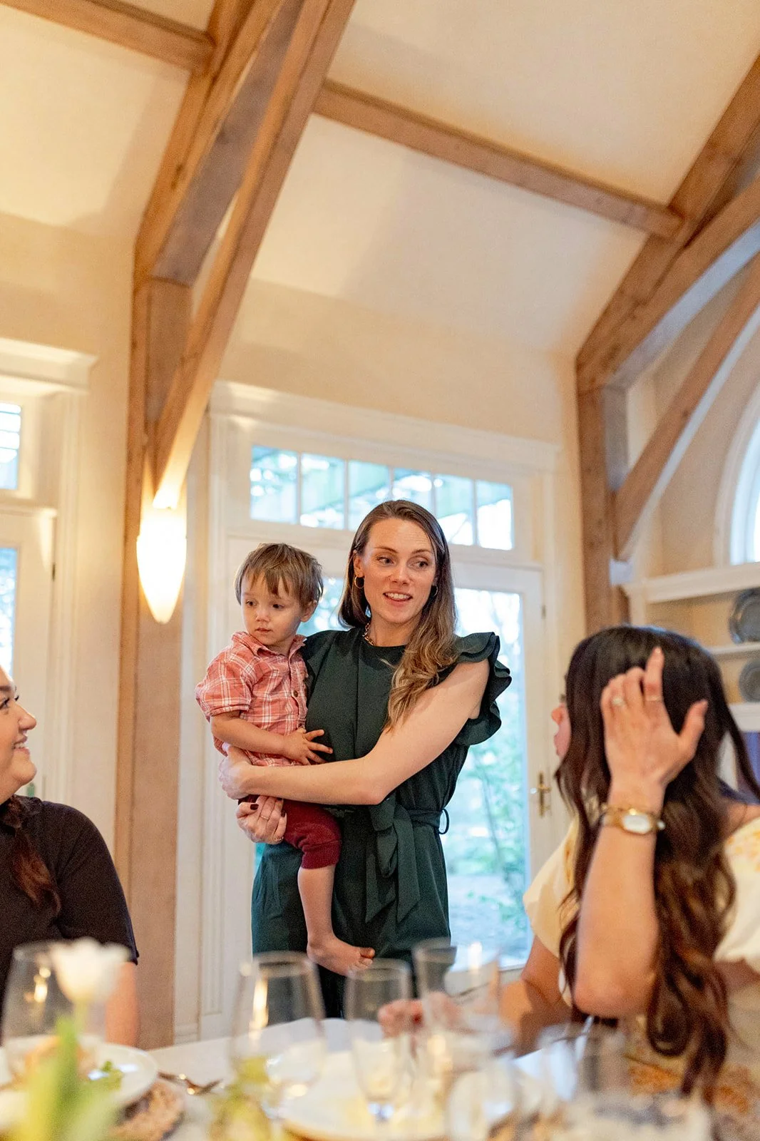 A woman holding a young boy at a social gathering or family dinner inside a room with large windows and wooden beams.