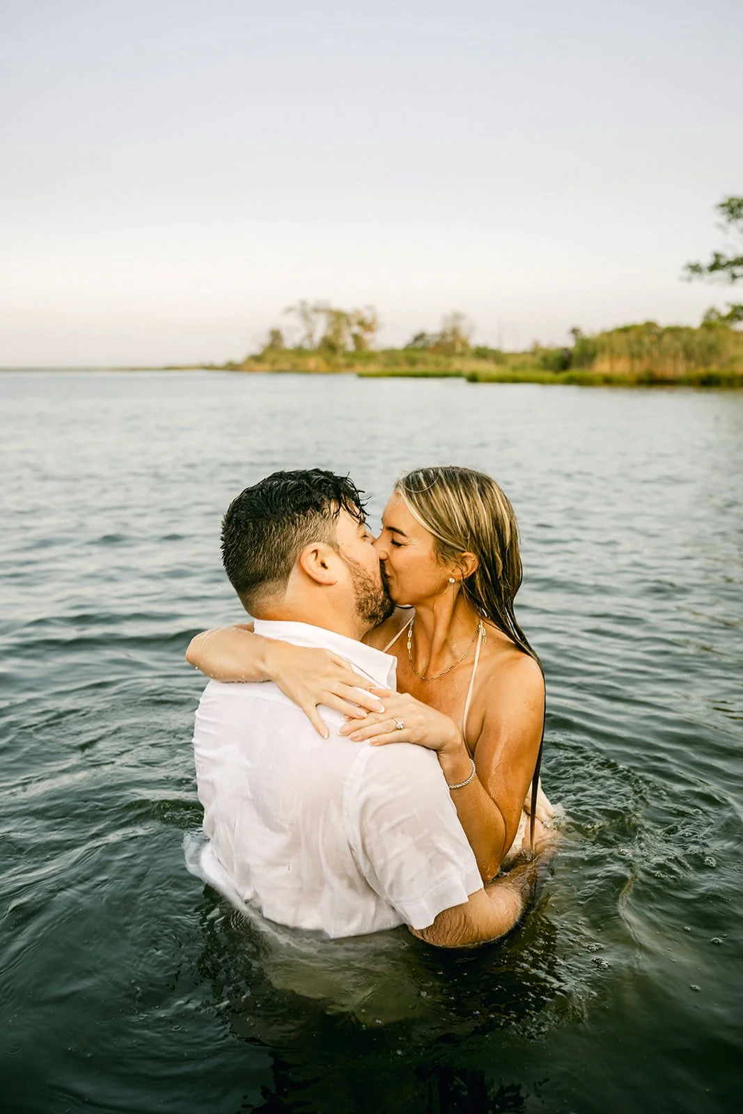 A couple kissing in a lake during sunset, with the woman wearing a dress and the man in a white shirt.