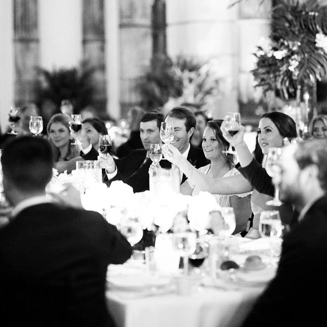 People at a banquet raising glasses in a toast, seated around a table with floral centerpiece and glasses of wine, in a formal setting.