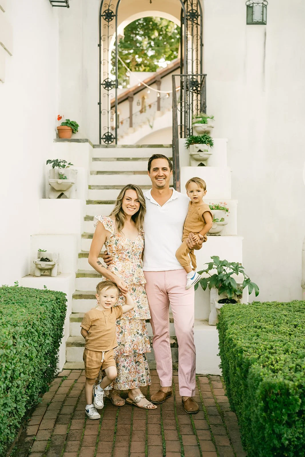 Family of four standing on a brick pathway in front of a white staircase with potted plants, smiling outdoors during daytime.