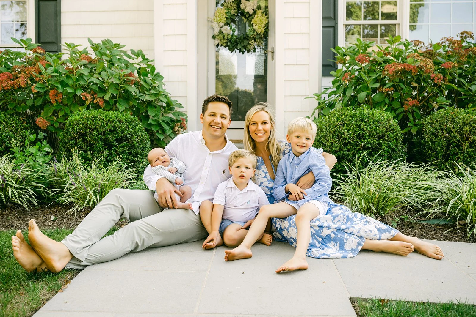 A happy family of five sitting on a concrete patio in front of their house, surrounded by lush green bushes and plants, with a white door decorated with a floral wreath in the background.