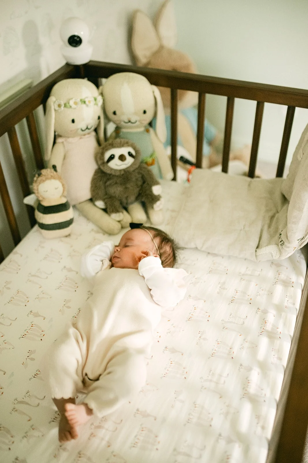 A sleeping baby in a white onesie lying in a crib with plush toys behind. The toys include rabbits, a sloth, a doll, and a character with a round head and black and white stripes.