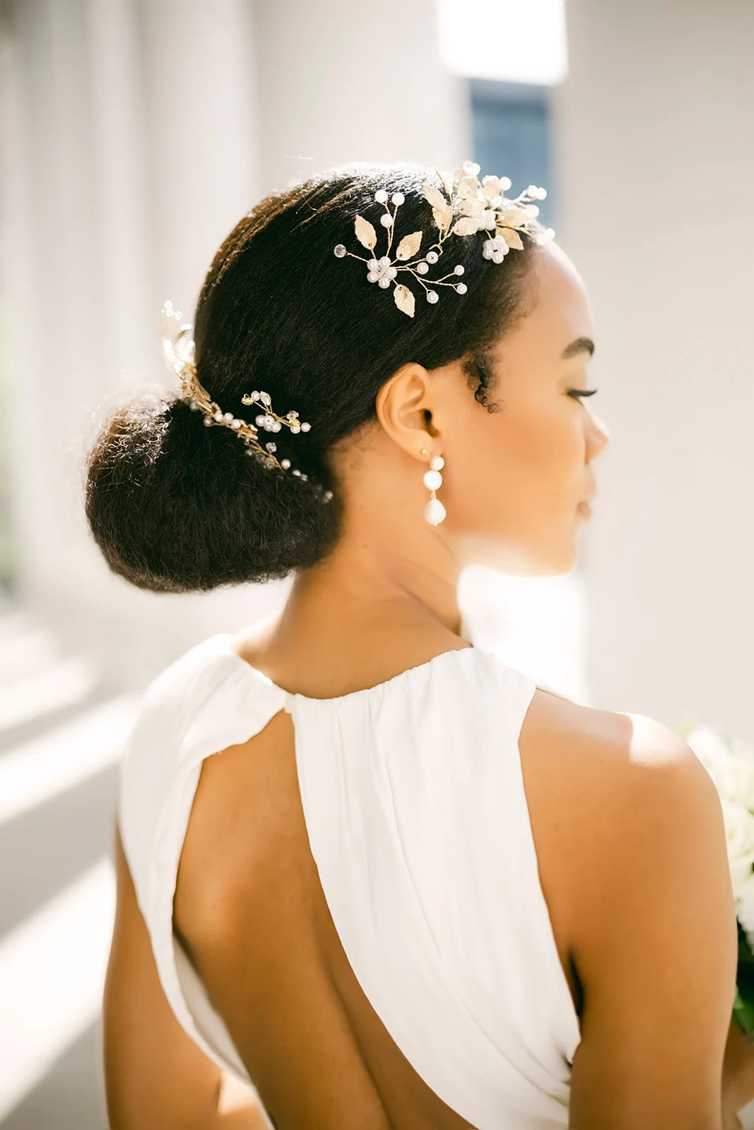 Close-up of a woman with dark hair styled in an elegant bun, adorned with a decorative hairpiece and pearl accents. She wears drop earrings and a white dress with open back details, standing in soft sunlight.