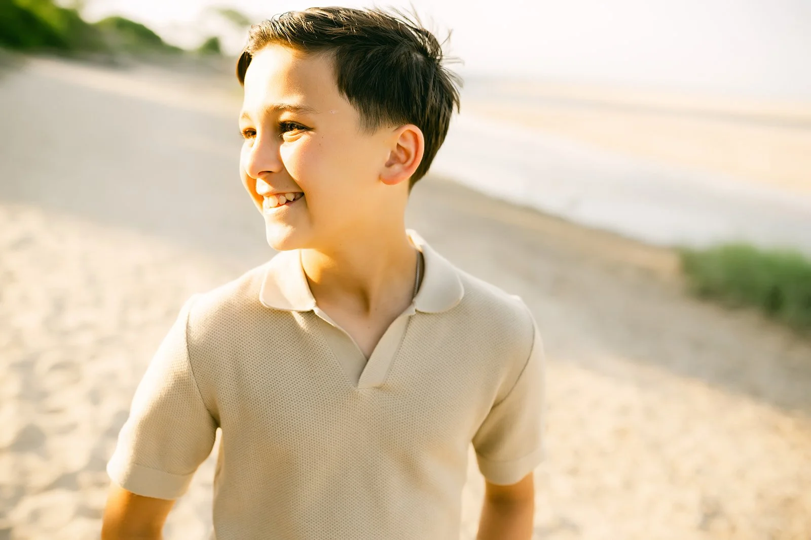 A young boy smiling and looking to his left on a beach during sunset, wearing a beige short-sleeve polo shirt.