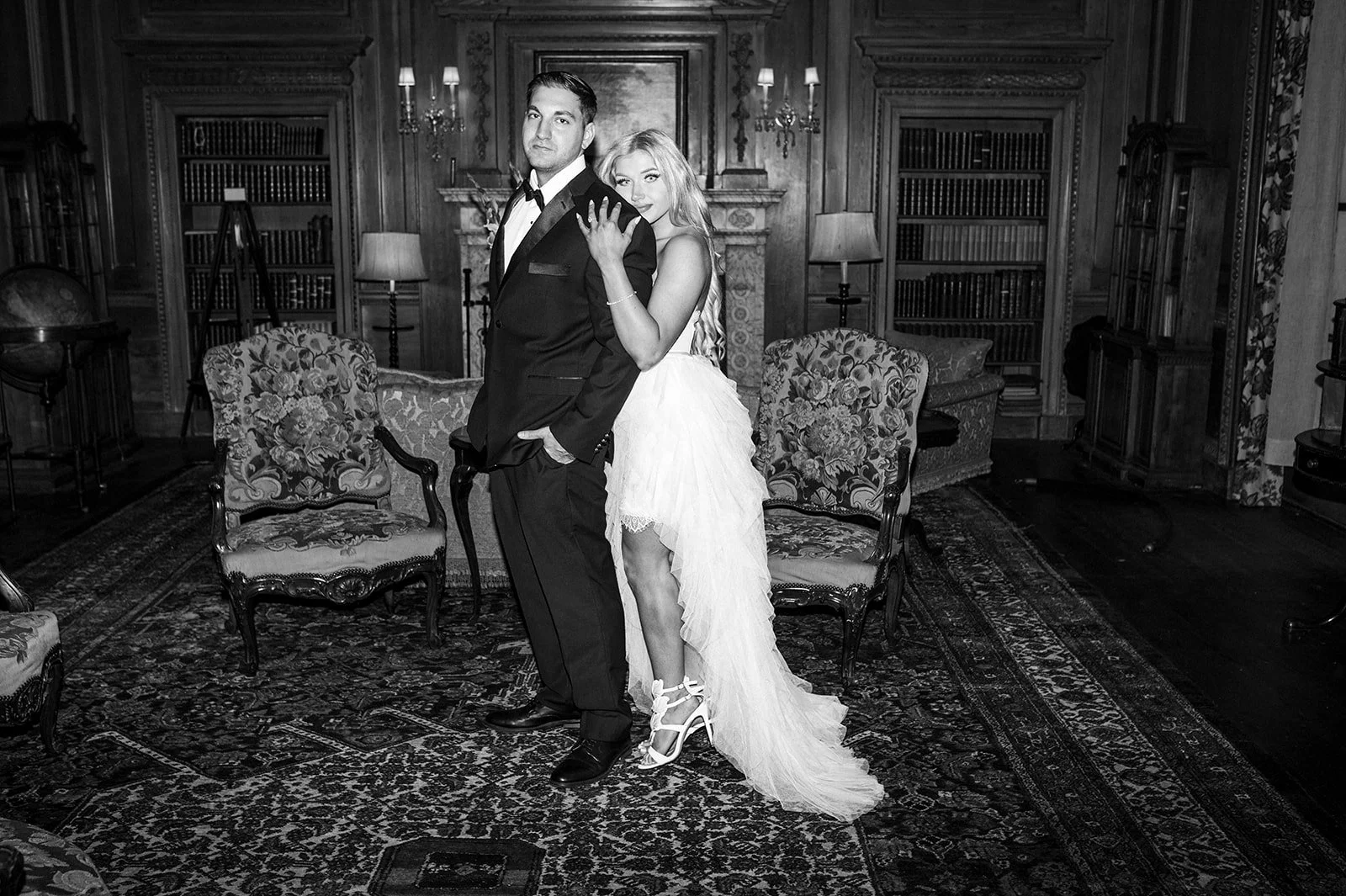 A couple in formal attire standing in a vintage library or study room with wooden walls, bookshelves, and ornate furniture.
