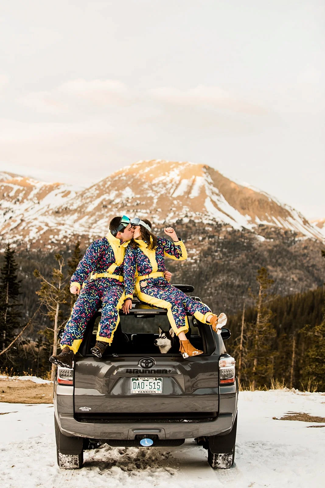 Two people in colorful ski suits sharing a kiss on top of a black Toyota SUV with a Siberian Husky in the back seat, against a mountain backdrop with snow and trees.