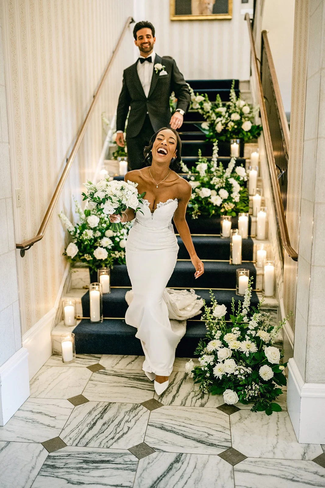A bride in a white wedding dress holding a bouquet, smiling and walking down a staircase behind a groom in a black tuxedo with a white shirt and bow tie, decorated with white flowers and candles.