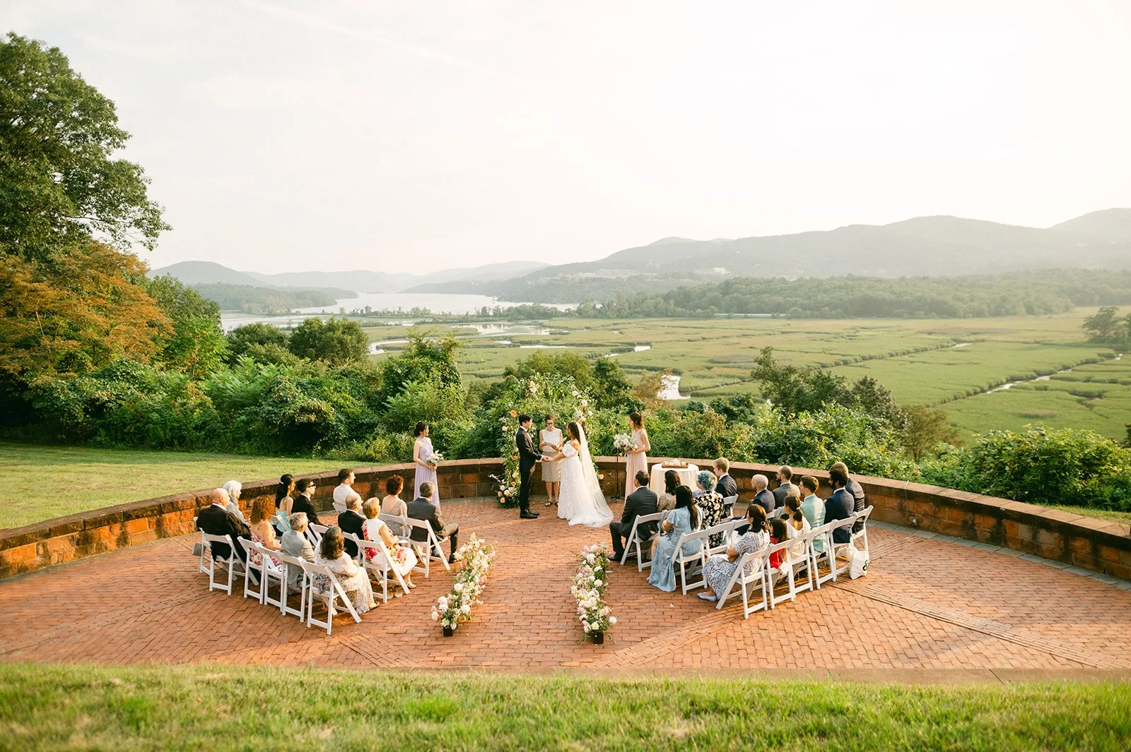 Outdoor wedding ceremony on a brick patio with scenic river and mountain view in the background, featuring a bride and groom exchanging vows surrounded by seated guests and officiants, decorated with flowers.