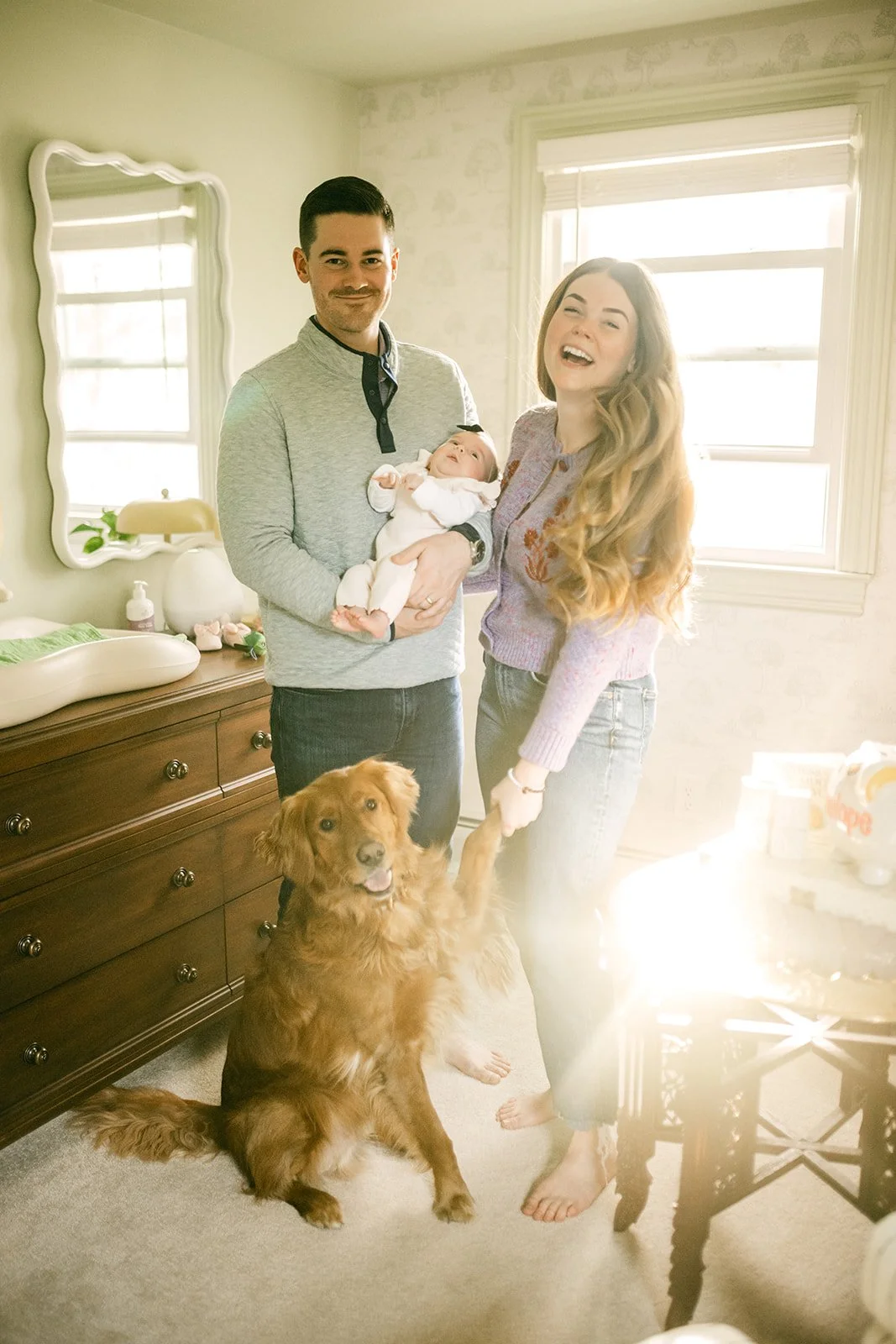 A happy family of three with their pet dog in a warmly lit bedroom. The man is holding a newborn baby, and the woman is happily holding hands with the dog. The sunlight streams in through the window.