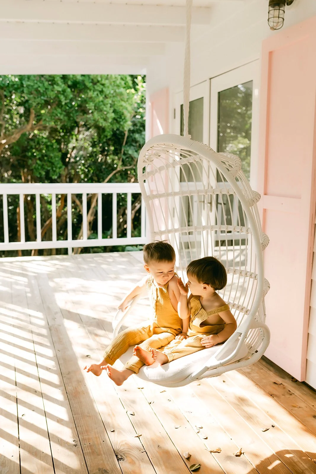 Two young children sitting on a white hanging bench on a porch, with one whispering to the other, surrounded by soft sunlight and green trees in the background.