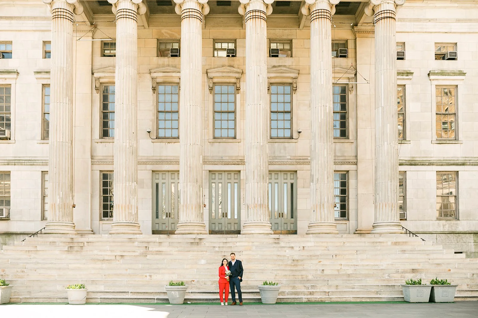A man and woman standing on steps in front of a large neoclassical building with tall columns and multiple windows, holding flowers.