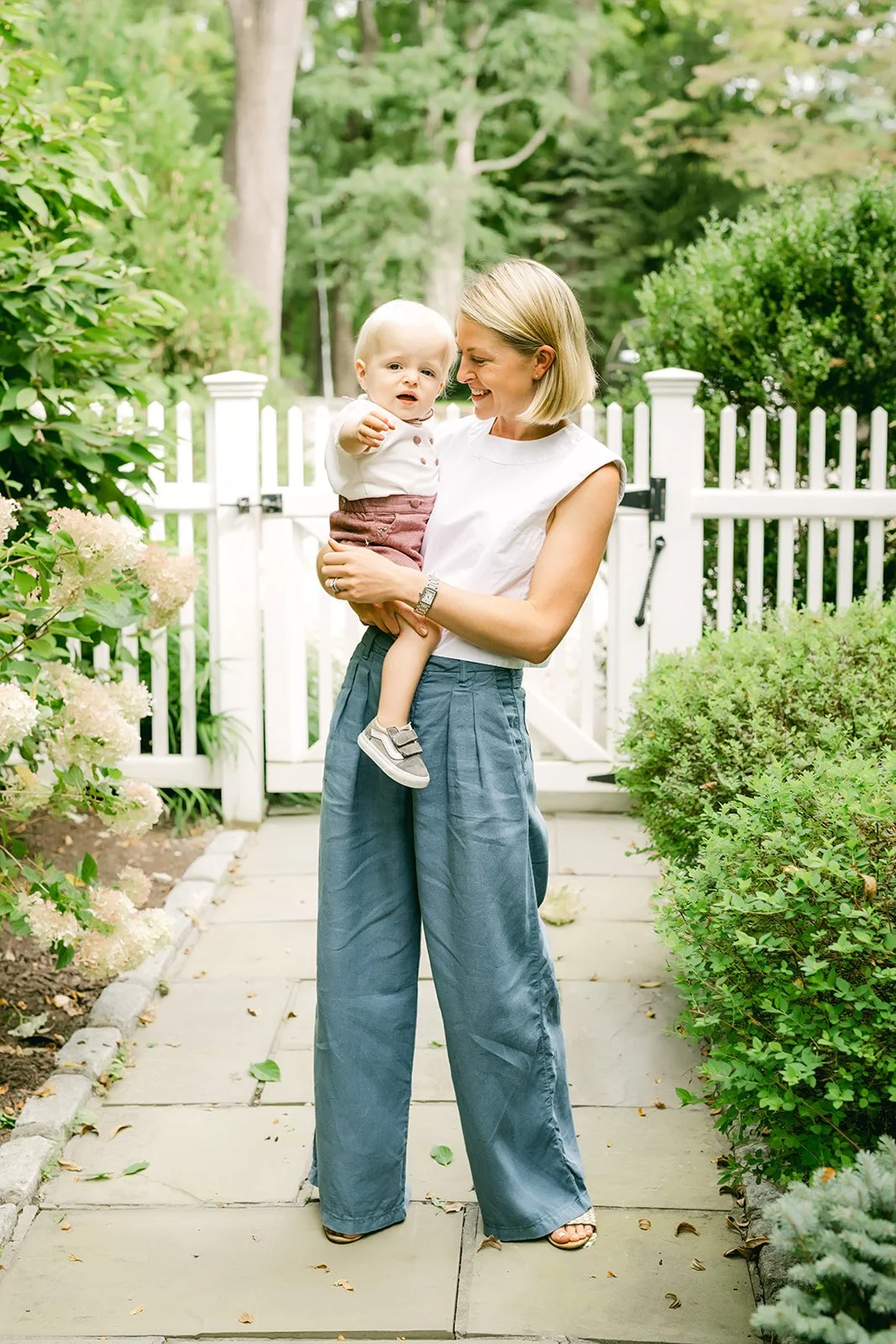 A woman holding a young child on a garden path with a white picket fence, greenery, and trees in the background.