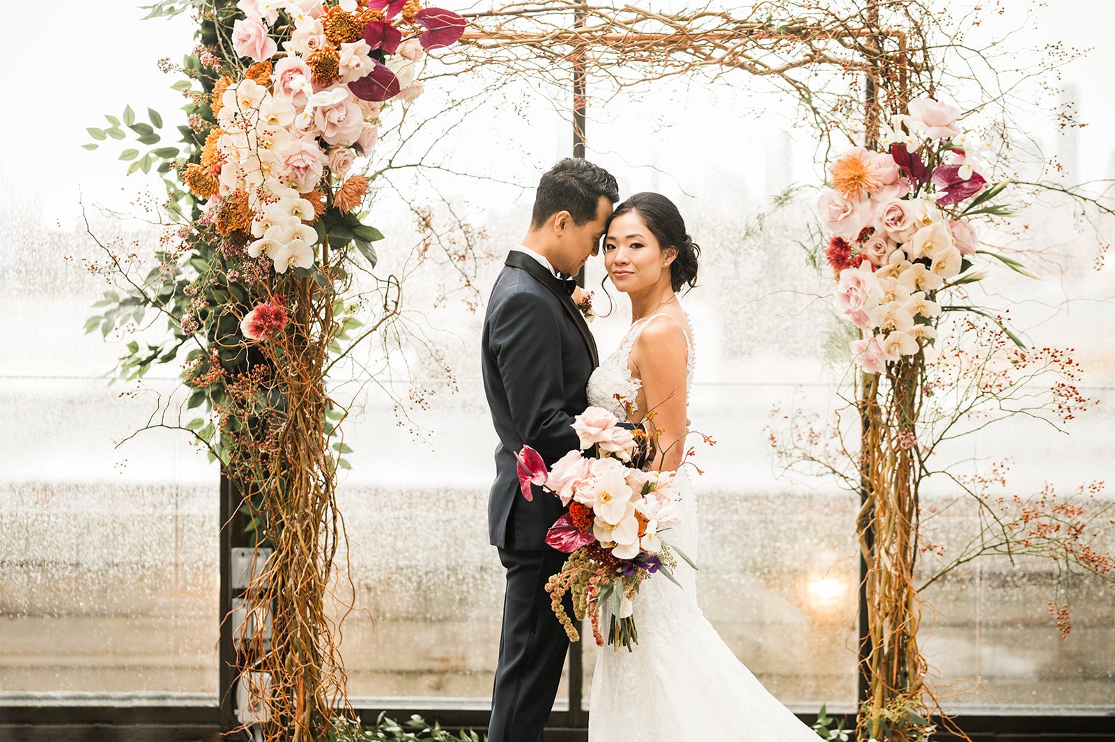 A bride and groom standing under a floral wedding arch, facing each other, with a glass window in the background