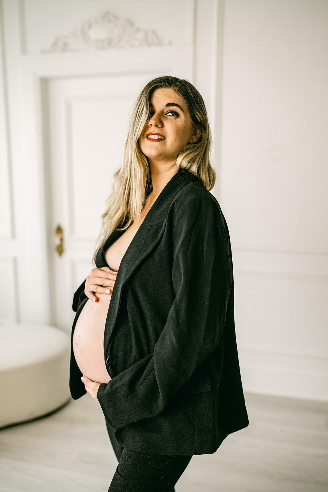 Pregnant woman with long blonde hair wearing a black blazer, standing in a room with white walls, touching her baby bump and looking at the camera.