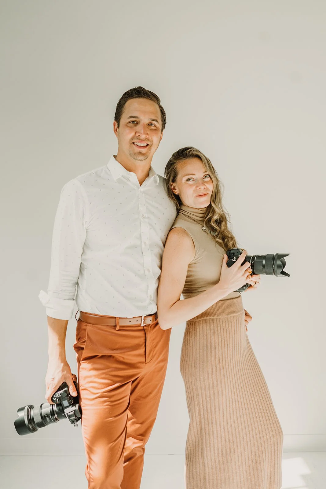 A man and woman stand together, holding professional cameras, smiling slightly, against a plain white background.