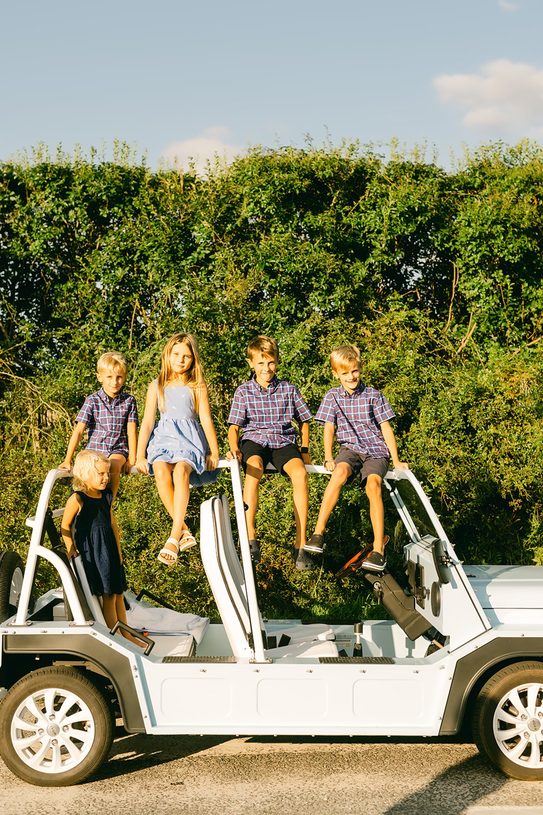 Five children, three boys and two girls, sitting on the open roof of a white off-road vehicle, parked outdoors in front of green bushes and blue sky.