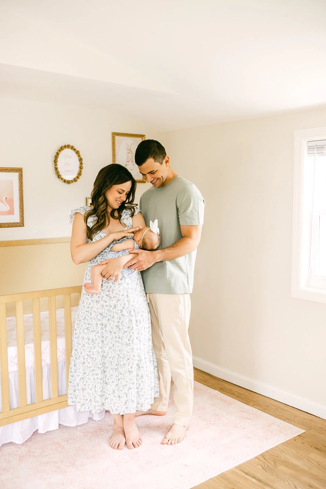 A couple stands in a bright room holding a newborn baby girl dressed in a blue outfit and white bow headband. The woman has long dark hair and is wearing a floral dress, while the man has short dark hair and is wearing a light green t-shirt and light-colored pants. They are smiling and looking at the baby.