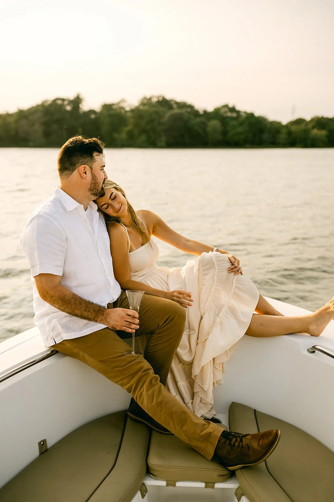 A couple sitting on a boat by the lake, with the woman resting her head on the man's shoulder, both holding drinks, during sunset.