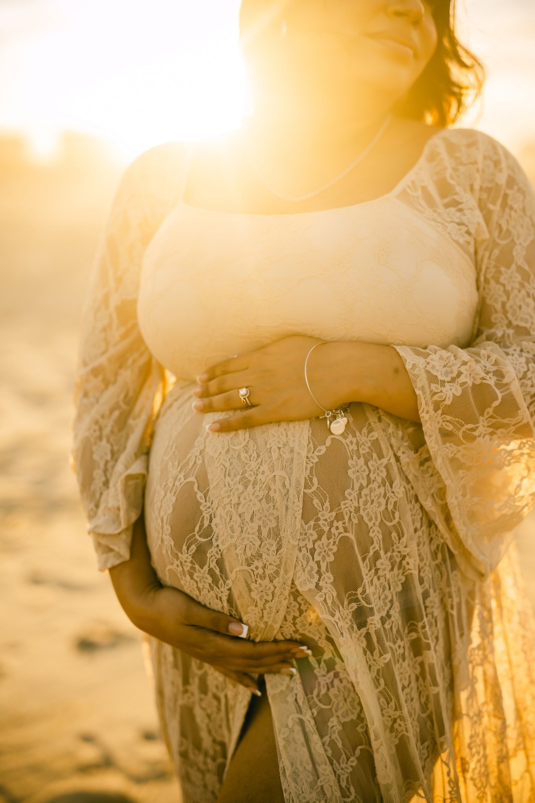 A pregnant woman in a sheer lace dress standing outdoors in the sunlight, holding her belly with one hand and wearing jewelry.