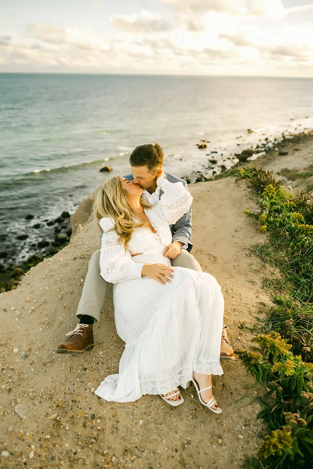 A couple sitting on a rocky ledge by the ocean, sharing a kiss, with a cloudy sky overhead.