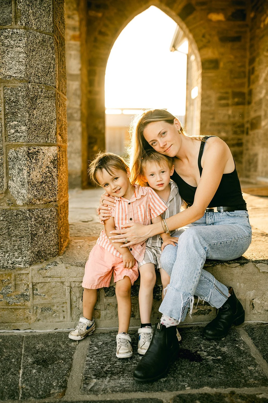A woman with two young children sitting on stone steps under an archway, with sunlight shining behind them.
