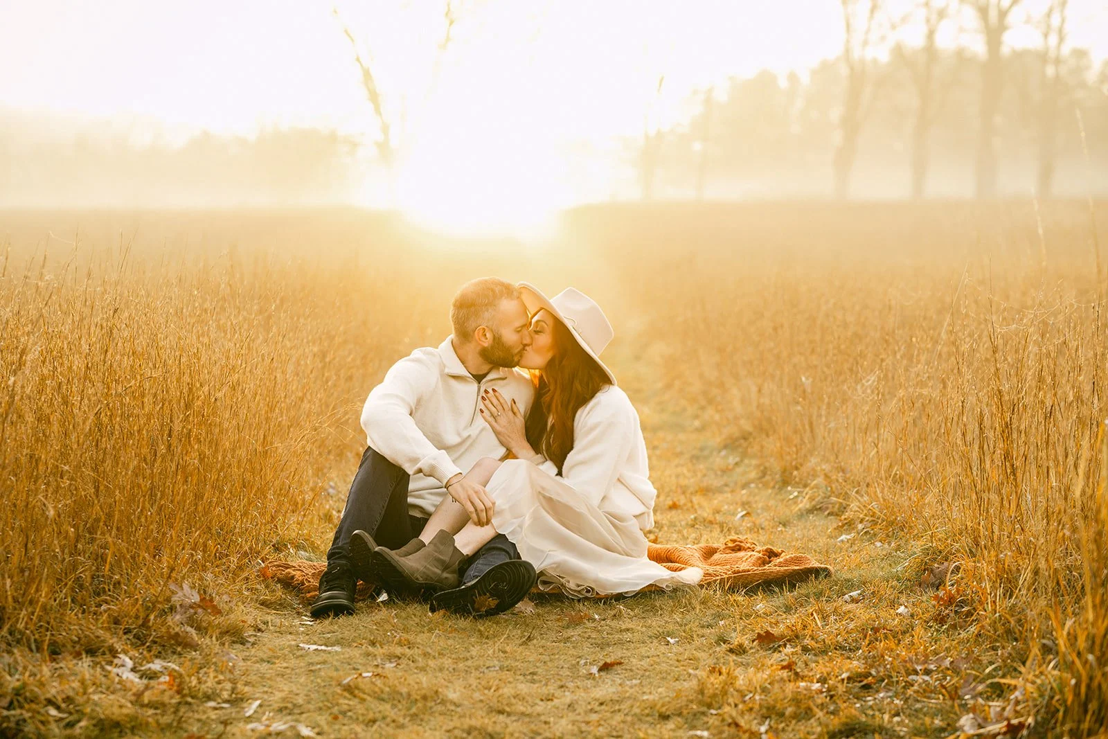 A couple sitting on a blanket in a field during sunset, sharing a kiss.