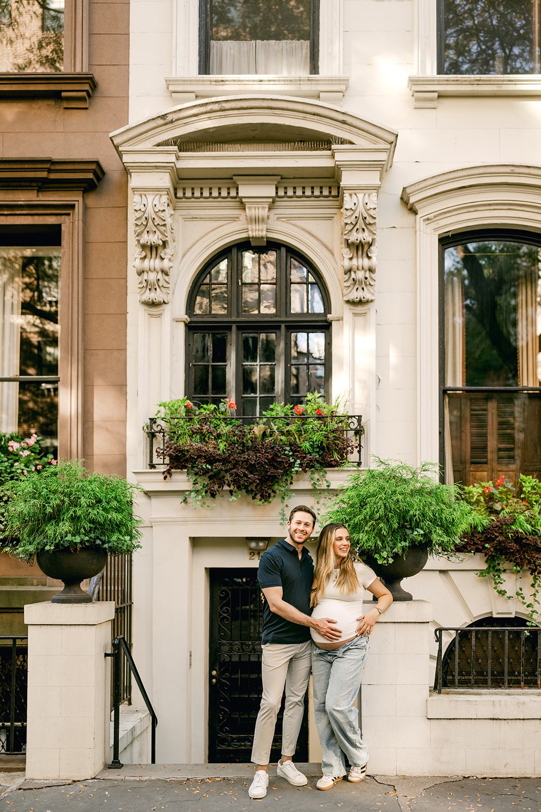 A couple standing in front of a brownstone or townhouse, with the woman showing her pregnant belly. They are smiling and embracing. The building has large windows with plants in front and decorative architecture around the windows.