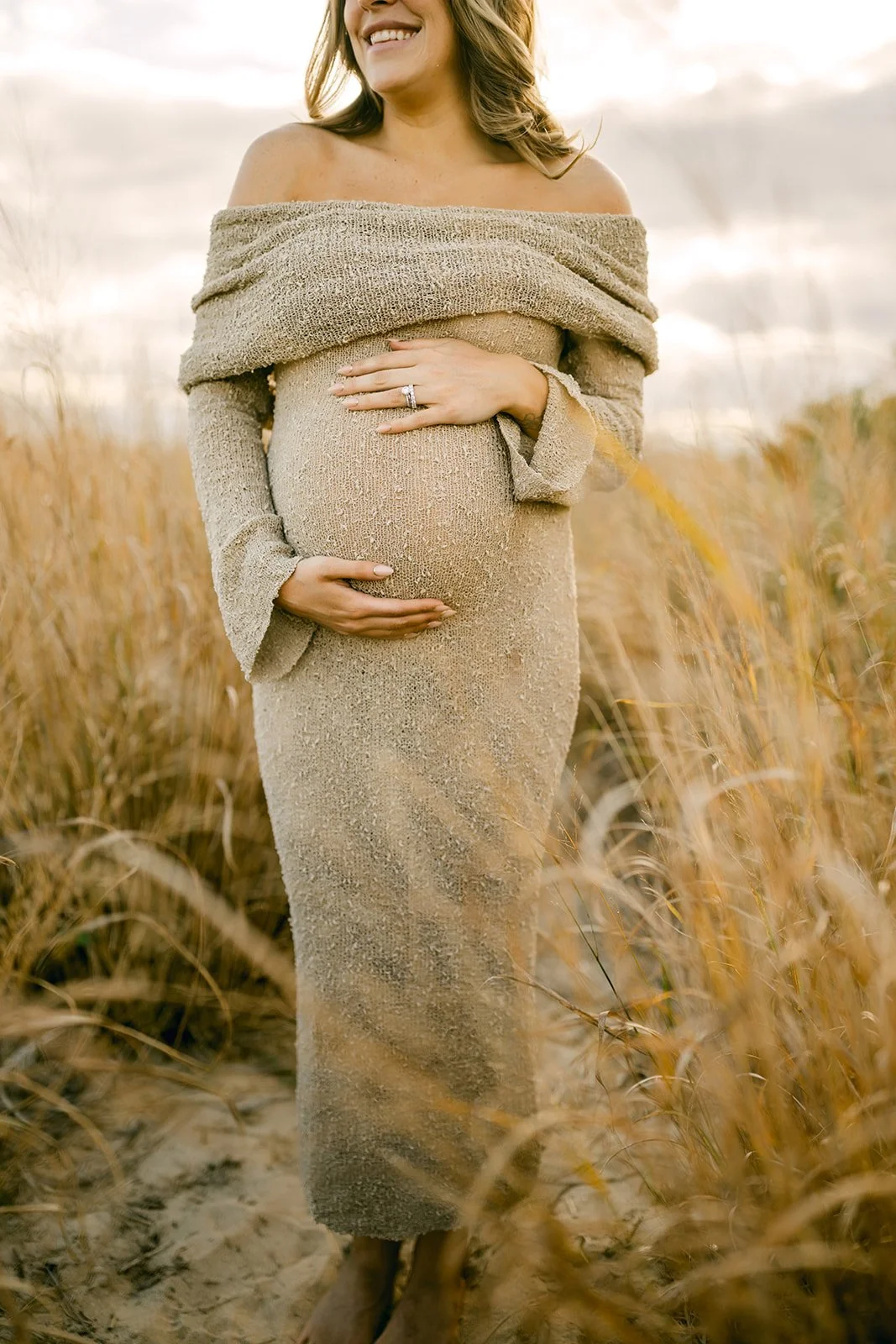 A pregnant woman standing in a field of tall grass, smiling, wearing a beige off-shoulder dress and holding her belly with her left hand, showing a ring.