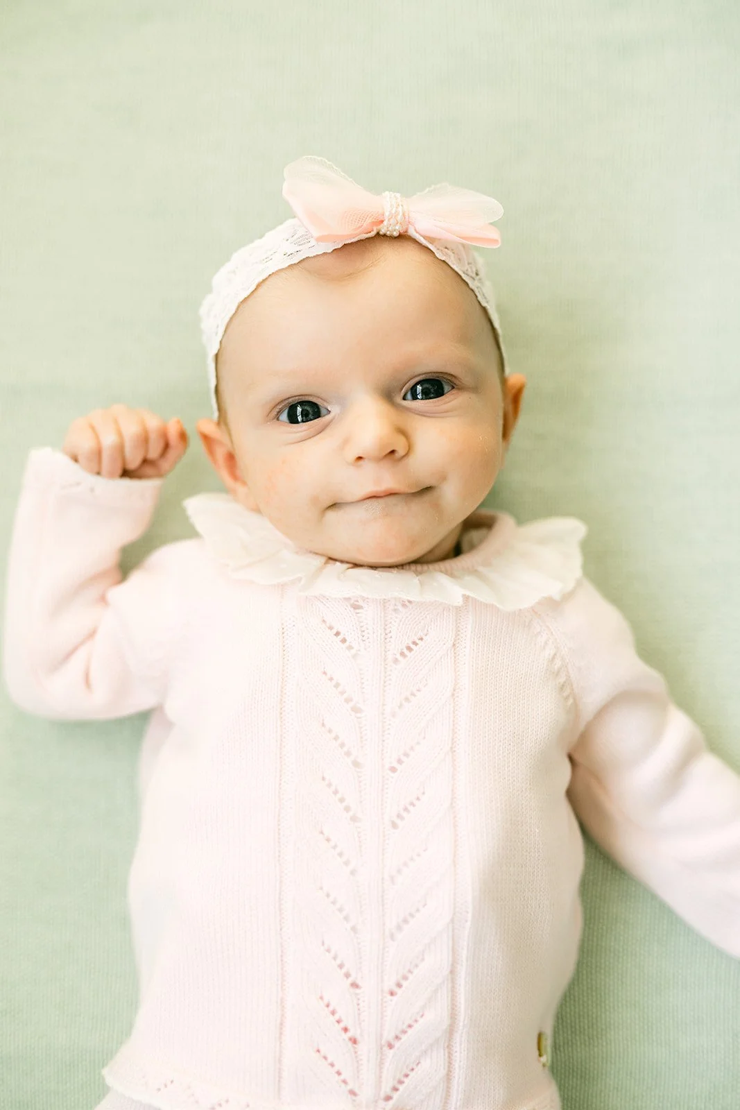 A baby girl with a pink bow headband lying on a light green surface, wearing a white sweater with a ruffled collar, smiling and raising her fist.