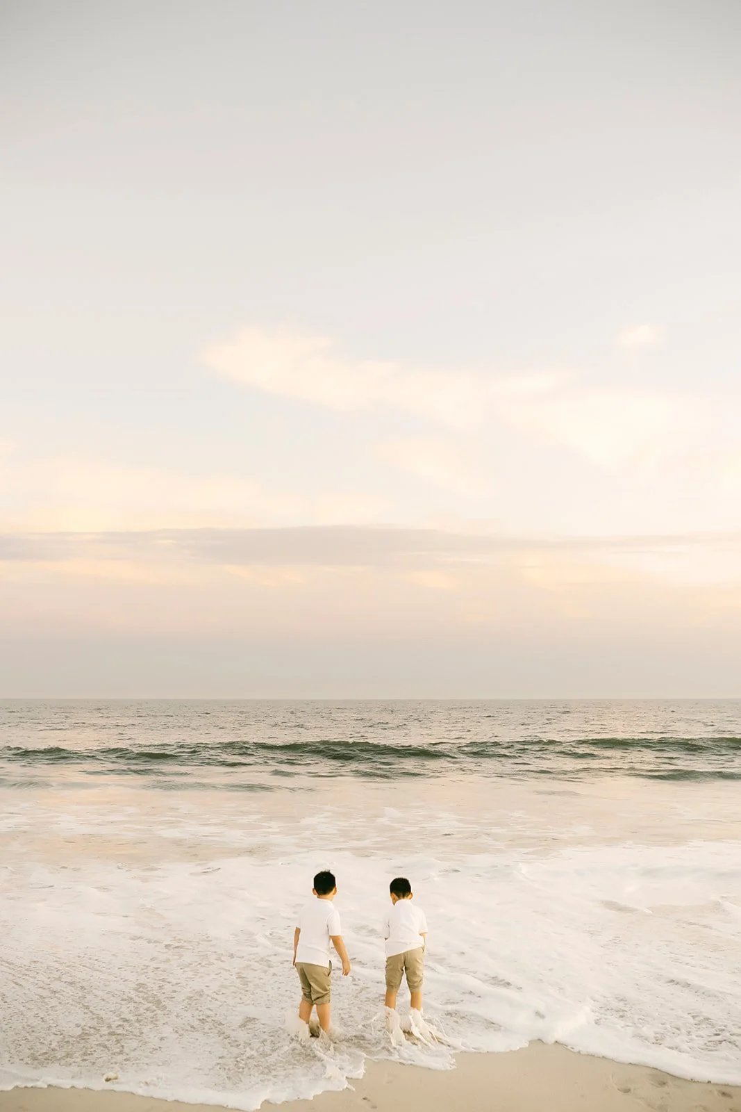 Two children standing at the edge of the ocean, facing the water, wearing white shirts and khaki shorts, with a cloudy sky above the horizon.